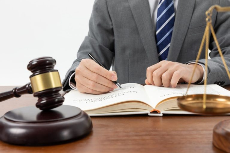 Hombre en traje firmando un documento con un martillo de juez y una balanza en la mesa.